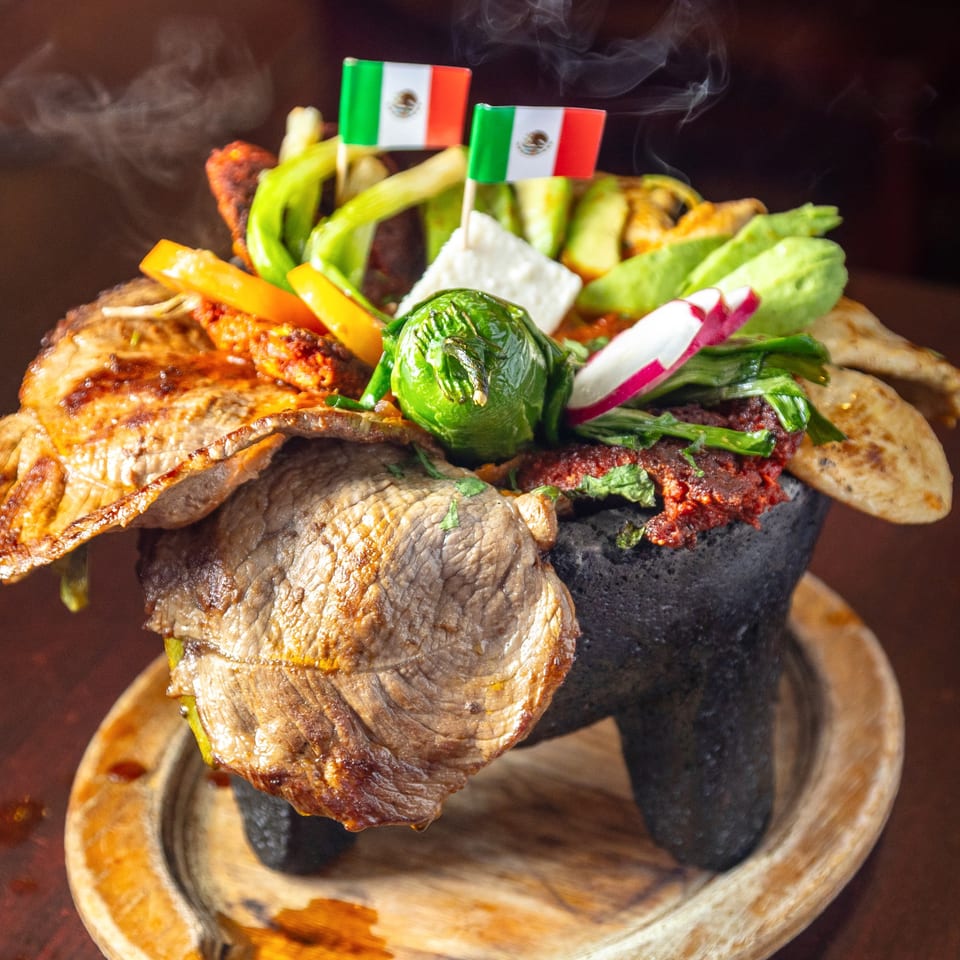 Steaming molcajete filled with grilled meats, vegetables, cheese, and two Mexican flags.