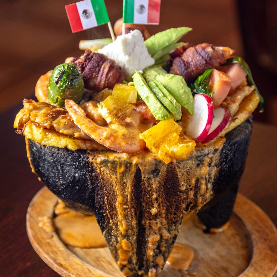 A molcajete overflowing with Mexican food, including meat, cheese, avocado, pineapple, and radishes.