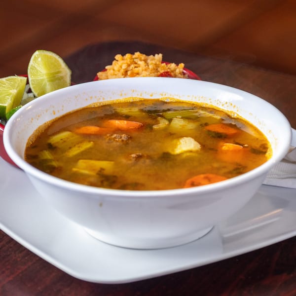 Hearty Mexican soup with carrots, celery, and meat, served with lime wedges and rice.