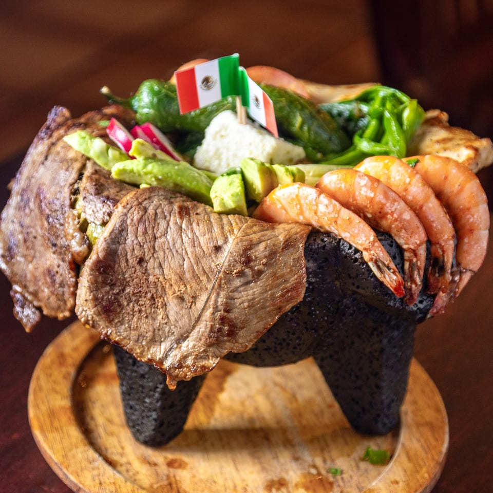 Molcajete dish with grilled steak, shrimp, avocado, radishes, and peppers, garnished with Mexican flags.